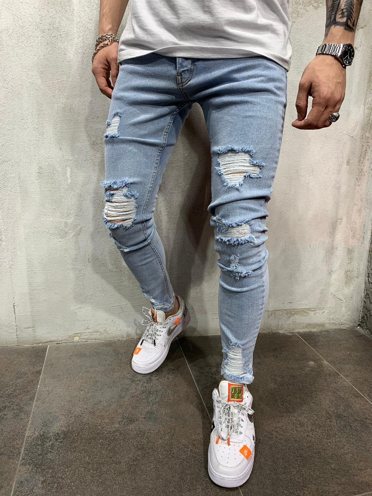 Light wash blue ripped skinny jeans worn by a model, paired with white sneakers and a white t-shirt, standing against a textured gray wall.