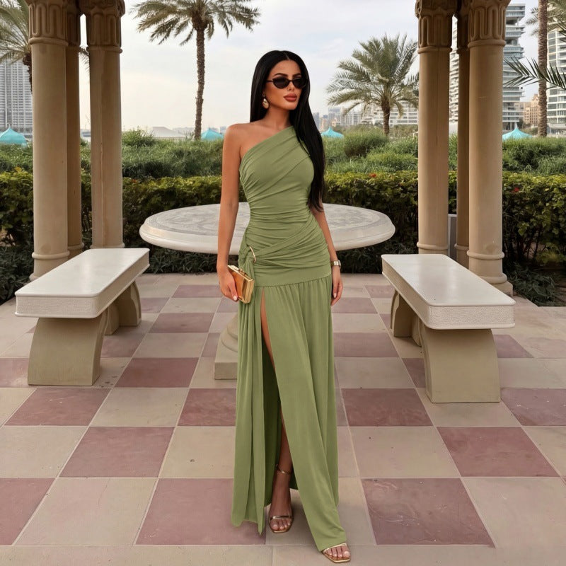 A woman stands in an outdoor terrace setting wearing a one-shoulder sage green maxi dress with a ruched bodice, thigh-high slit, and flowing skirt. She is holding a small gold clutch and wearing gold sandals.