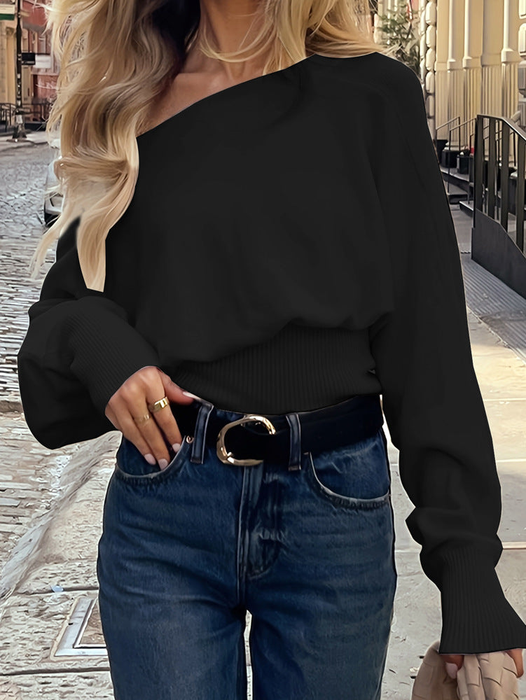 A woman wearing a black long-sleeve off-the-shoulder knit top tucked into blue jeans with a black belt, standing on a cobblestone street.