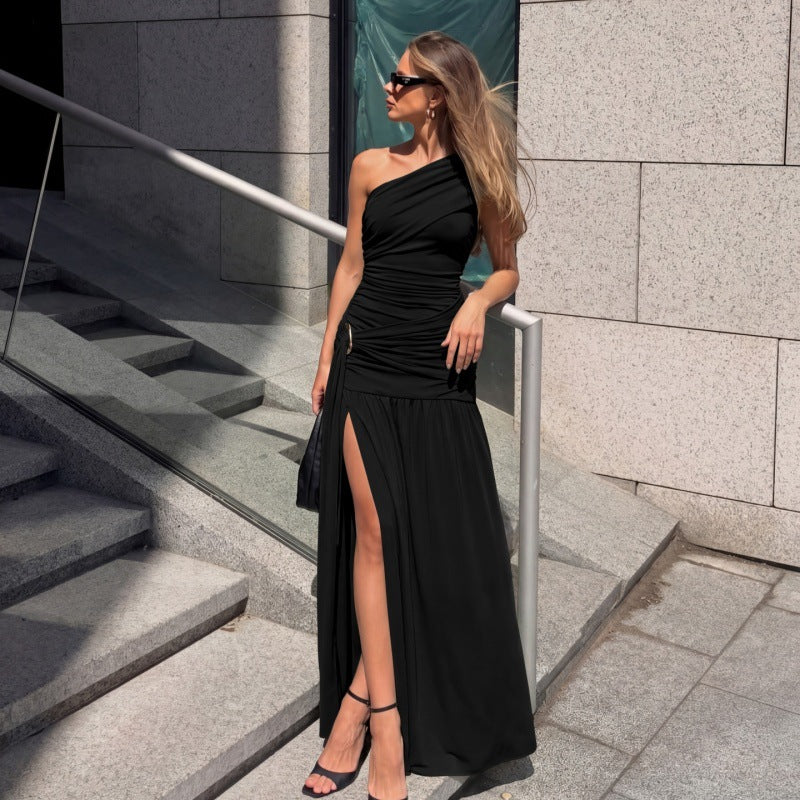A woman wearing a black one-shoulder maxi dress with a high thigh slit, standing on outdoor stone steps next to a glass railing.