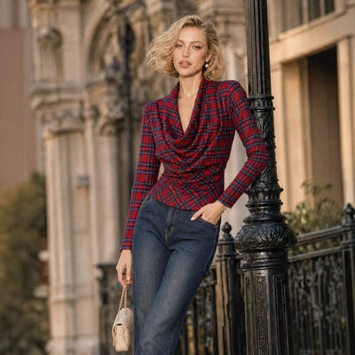 A woman stands outdoors leaning against a black metal post, wearing a long-sleeve red and navy plaid wrap-style top with dark blue jeans and holding a small beige handbag.