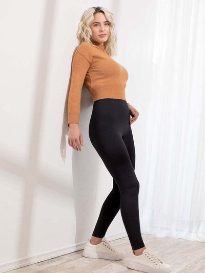 A woman posing in a room wearing a cropped, long-sleeve mustard yellow knit top, black leggings, and white sneakers.