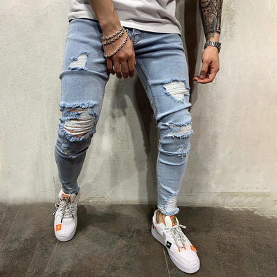 A person wearing light-wash distressed skinny jeans with ripped knees and frayed hems, paired with white sneakers, standing against a textured concrete wall.