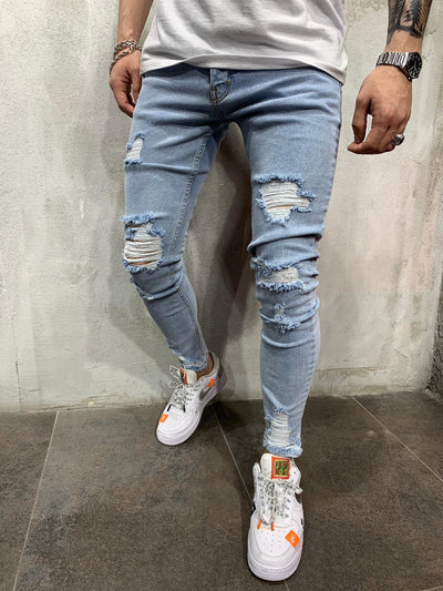 Light wash blue ripped skinny jeans worn by a model, paired with white sneakers and a white t-shirt, standing against a textured gray wall.
