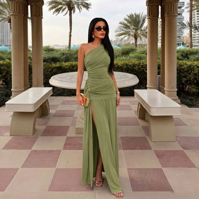 A woman stands in an outdoor terrace setting wearing a one-shoulder sage green maxi dress with a ruched bodice, thigh-high slit, and flowing skirt. She is holding a small gold clutch and wearing gold sandals.