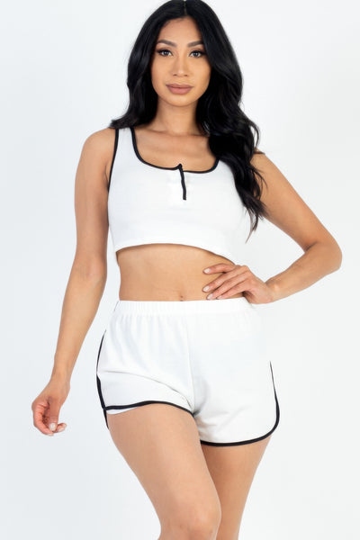 A female model wearing a white sleeveless tank top with button front and contrast trim, paired with matching shorts. The model is standing against a plain background.