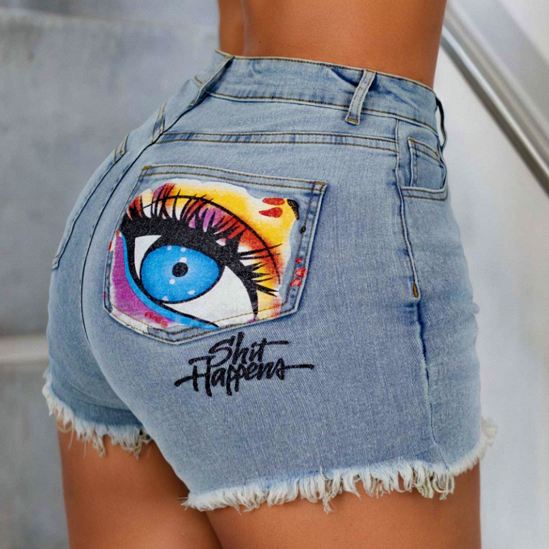 Light wash denim cut-off shorts with a large colorful eye graphic and the text 'Shit Happens' on the back pocket, shown on a model.