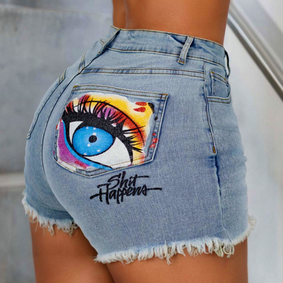 Light wash denim cut-off shorts with a large colorful eye graphic and the text 'Shit Happens' on the back pocket, shown on a model.