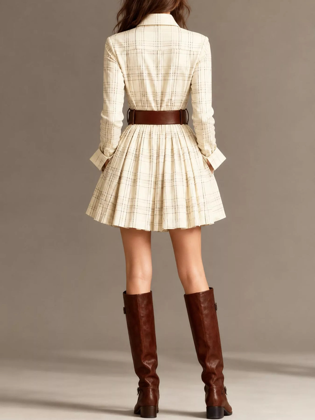 A model shown from the back wearing a cream and tan plaid mini dress with a pleated skirt, a brown belt, and brown knee-high leather boots, set against a solid taupe studio background.