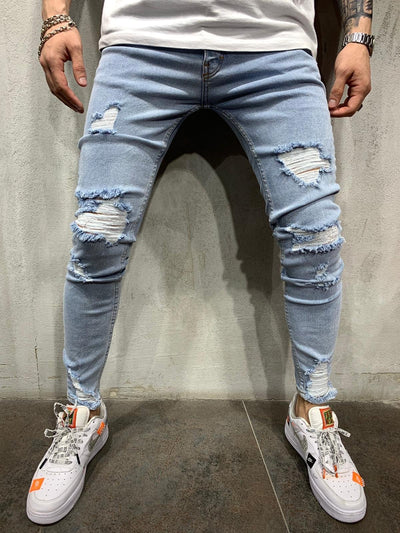 Light wash blue ripped skinny jeans worn by a model, paired with a white t-shirt and white sneakers with orange accents, standing against a gray concrete wall.