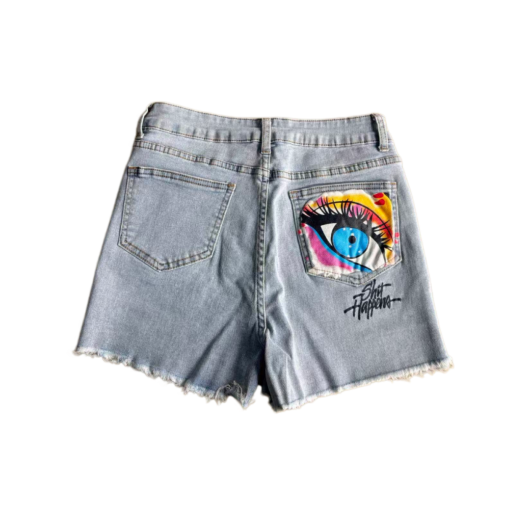 Light wash denim shorts with a distressed frayed hem, featuring a colorful graphic print of an eye on the back right pocket and a black script logo on the lower back pocket.