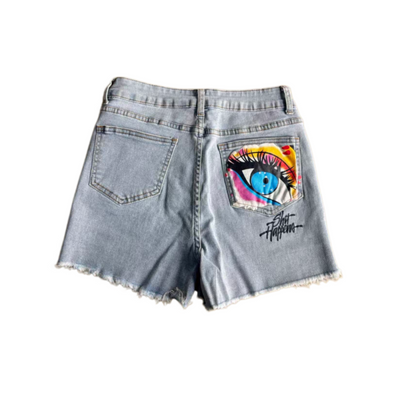Light wash denim shorts with a distressed frayed hem, featuring a colorful graphic print of an eye on the back right pocket and a black script logo on the lower back pocket.