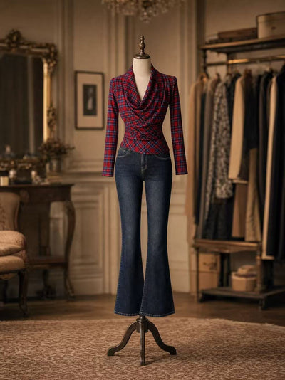 A mannequin dressed in a red and blue plaid cowl-neck long-sleeve top paired with dark blue flared jeans, displayed in an elegant vintage-style room with clothing racks and warm lighting.